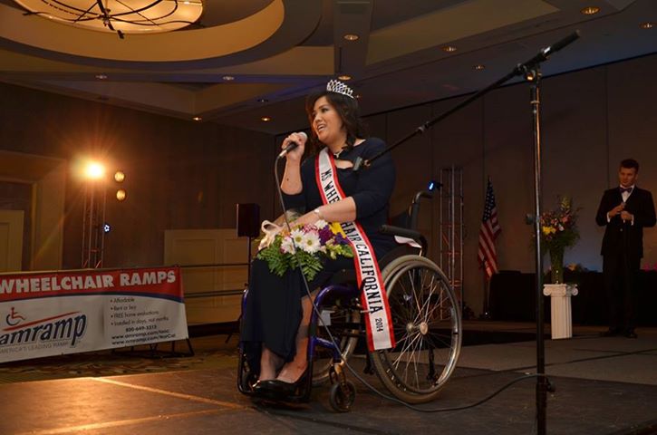 Theresa De Vera in wheelchair holding a microphone at an inside event with a crown and a sash reading "Ms Wheelchair California 2014"