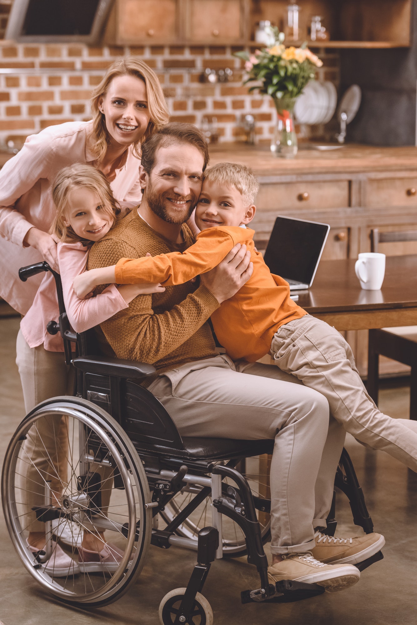 happy family with two kids and father in wheelchair hugging and smiling at camera at home
