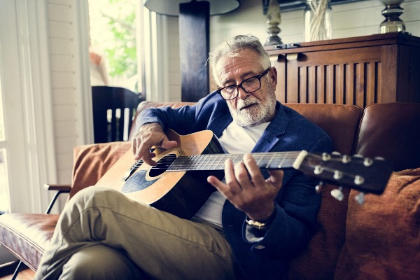Elderly an who is aging in place plays guitar from his couch at home.