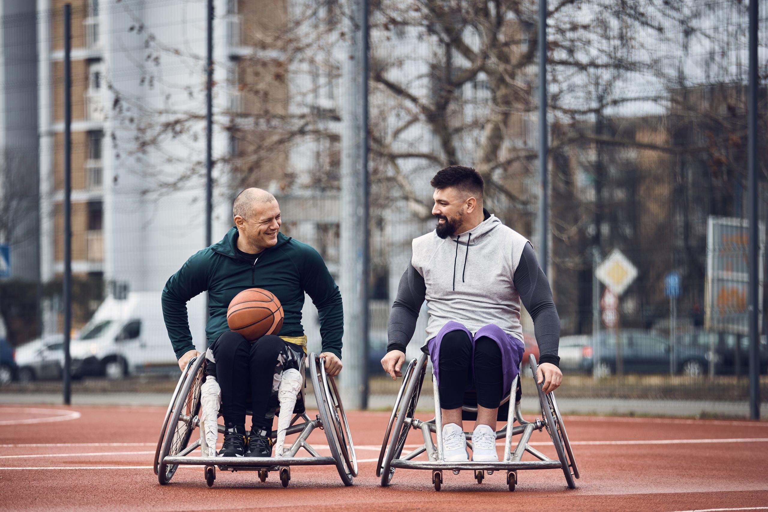 Two mean in wheelchairs laugh together as they prepare for a game of wheelchair basketball together.
