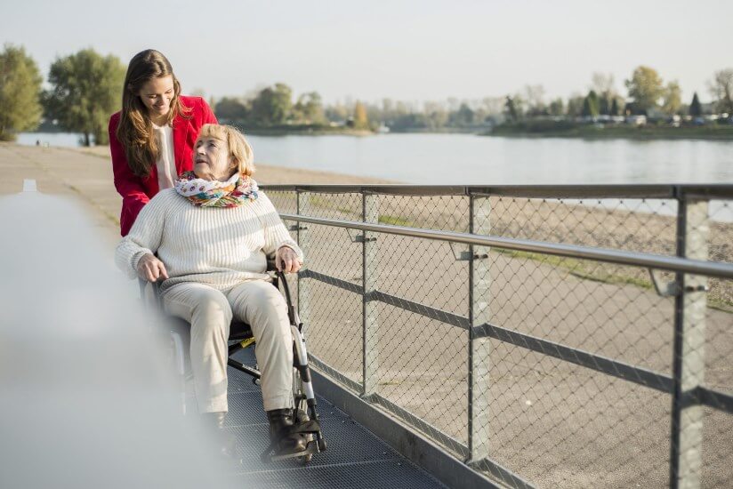 Woman in a red jacket walks a woman in a wheelchair up a modular wheelchair ramp near a pond