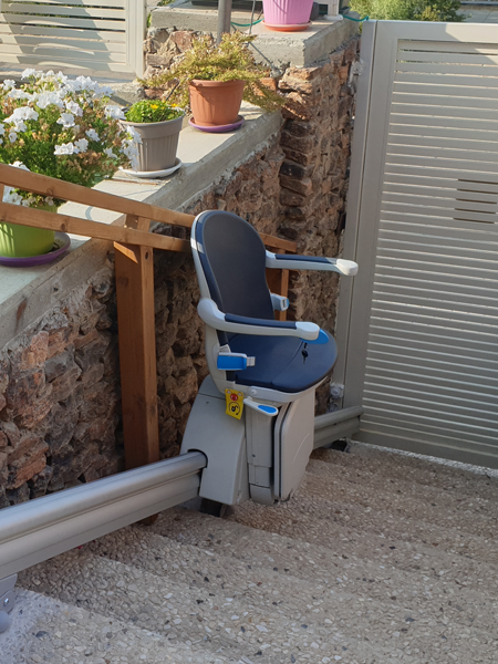 An outdoor stairlift is shown on a set of stairs leading to a house's entrance. The lift is designed to carry individuals with limited mobility up and down stairs safely and comfortably. It features a sturdy seat, armrests, and footrest, as well as safety sensors and a durable weather-resistant construction to withstand outdoor elements.