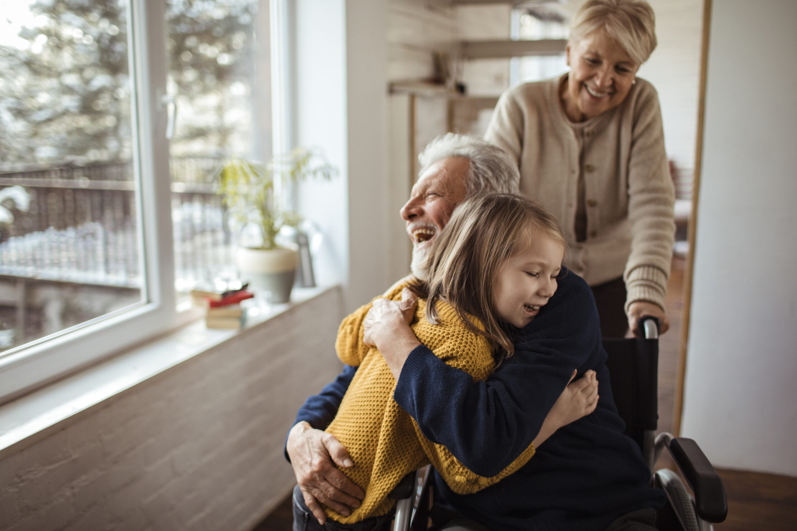 An older man in a wheelchair hugs his granddaughter. His wife stands behind him, pushing the chair.