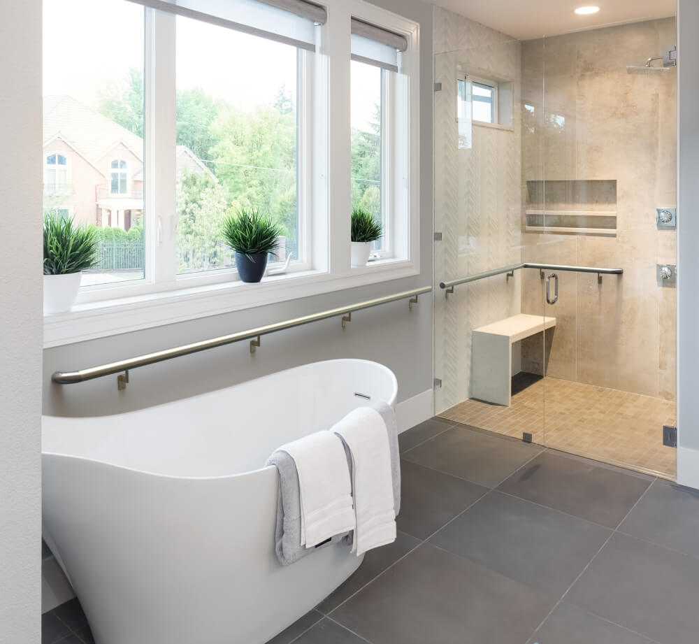 A white bathtub in a bathroom with a gray floor. Windows are above the bathroom looking out on some trees. There is a shower with a bench in the background.