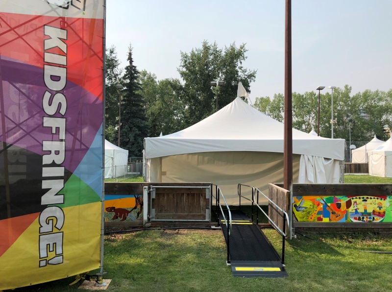 A view of a white festival tent with a ramp leading from grass up to a raised area.