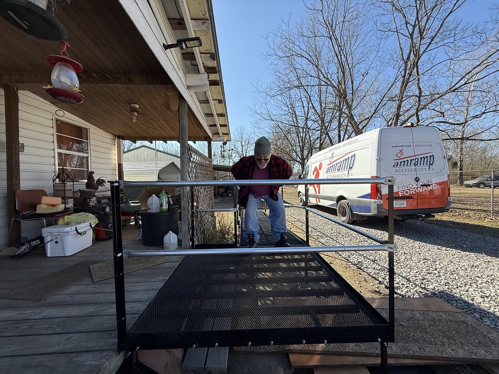 A man going up a steel wheelchair ramp with an Amramp work van in the background on a sunny day