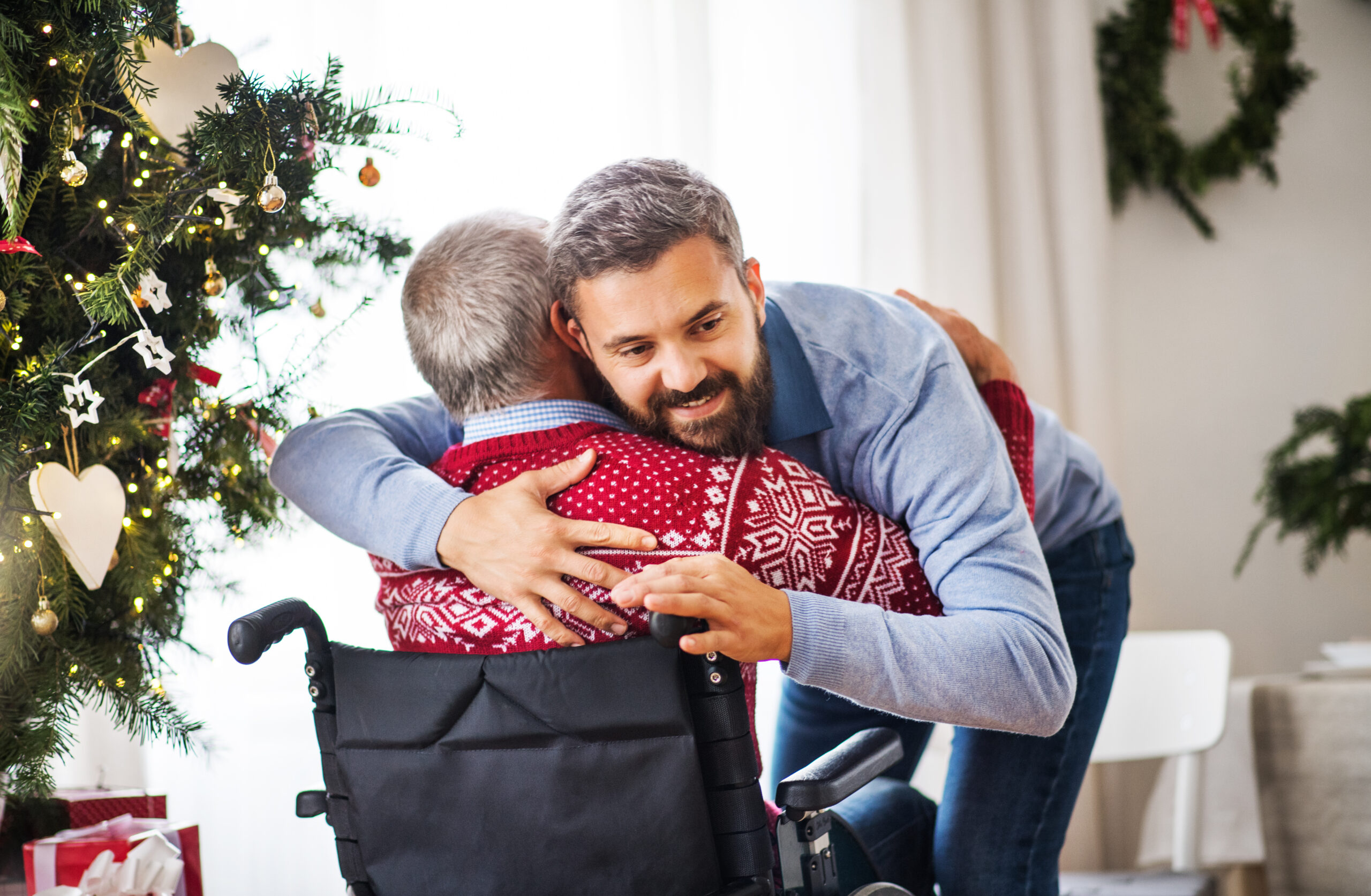 Man hugs his senior father in a wheelchair inside their home during the holidays.