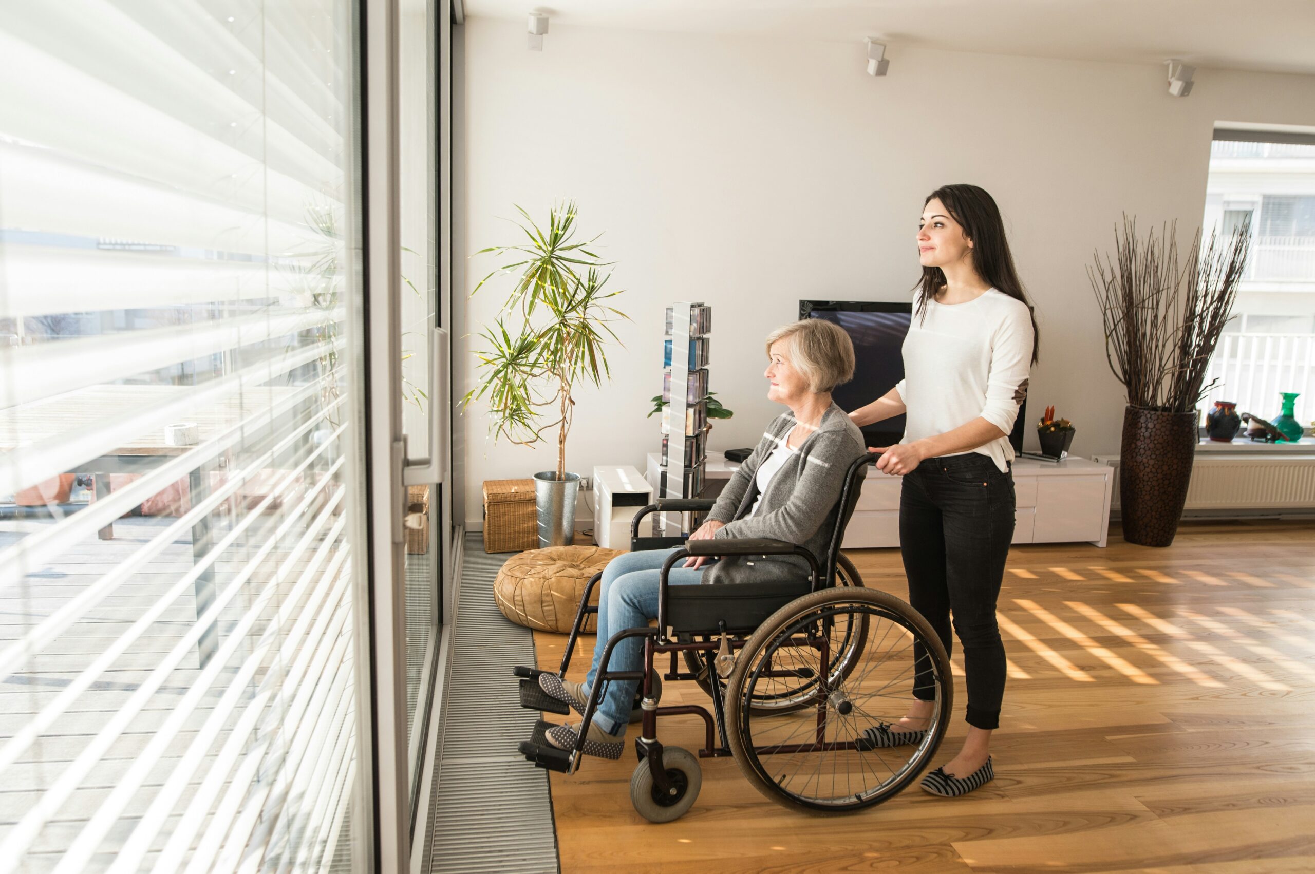 Older adult using a wheelchair indoors with assistance from a caregiver.