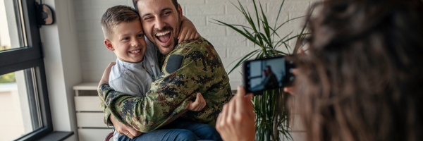 A U.S. Servicemember and Veteran hugs his young child while his wife takes a photo