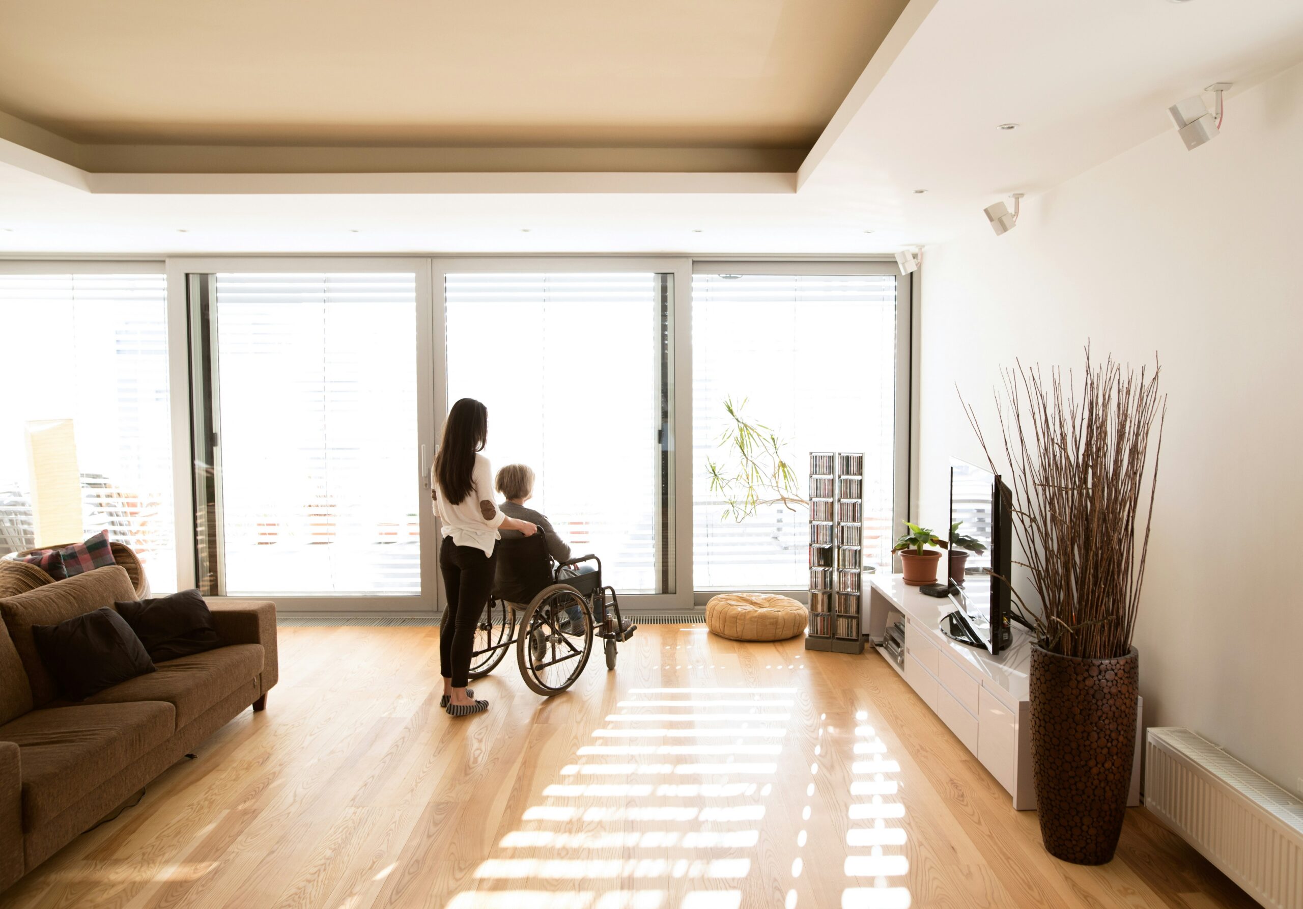 Caregiver standing beside an older adult using a wheelchair in a bright living room.