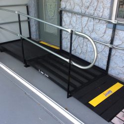 Short steel wheelchair ramp with handrails and yellow transition strips leading to a front door beside a stone exterior wall.