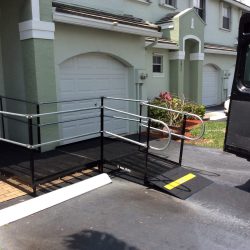 L-shaped steel wheelchair ramp with handrails connecting a driveway to the front entrance of a light green townhome.