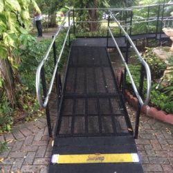 Steel wheelchair ramp with handrails rising from a brick walkway to a turning platform amid lush landscaping.