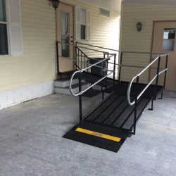 Steel wheelchair ramp with handrails and a turning platform leading from a covered concrete area to a raised home entrance.