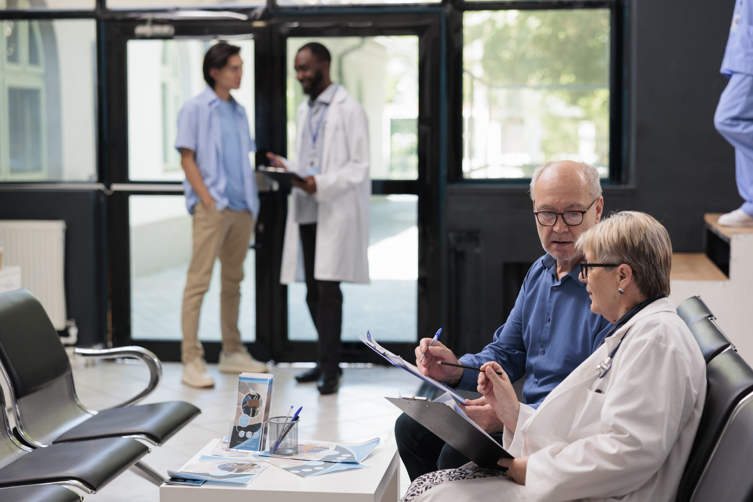 Senior doctor sitting on chair in hospital reception explaining disease diagnosis to elderly patient discussing health care treatment. People waiting to start examination during checkup visit