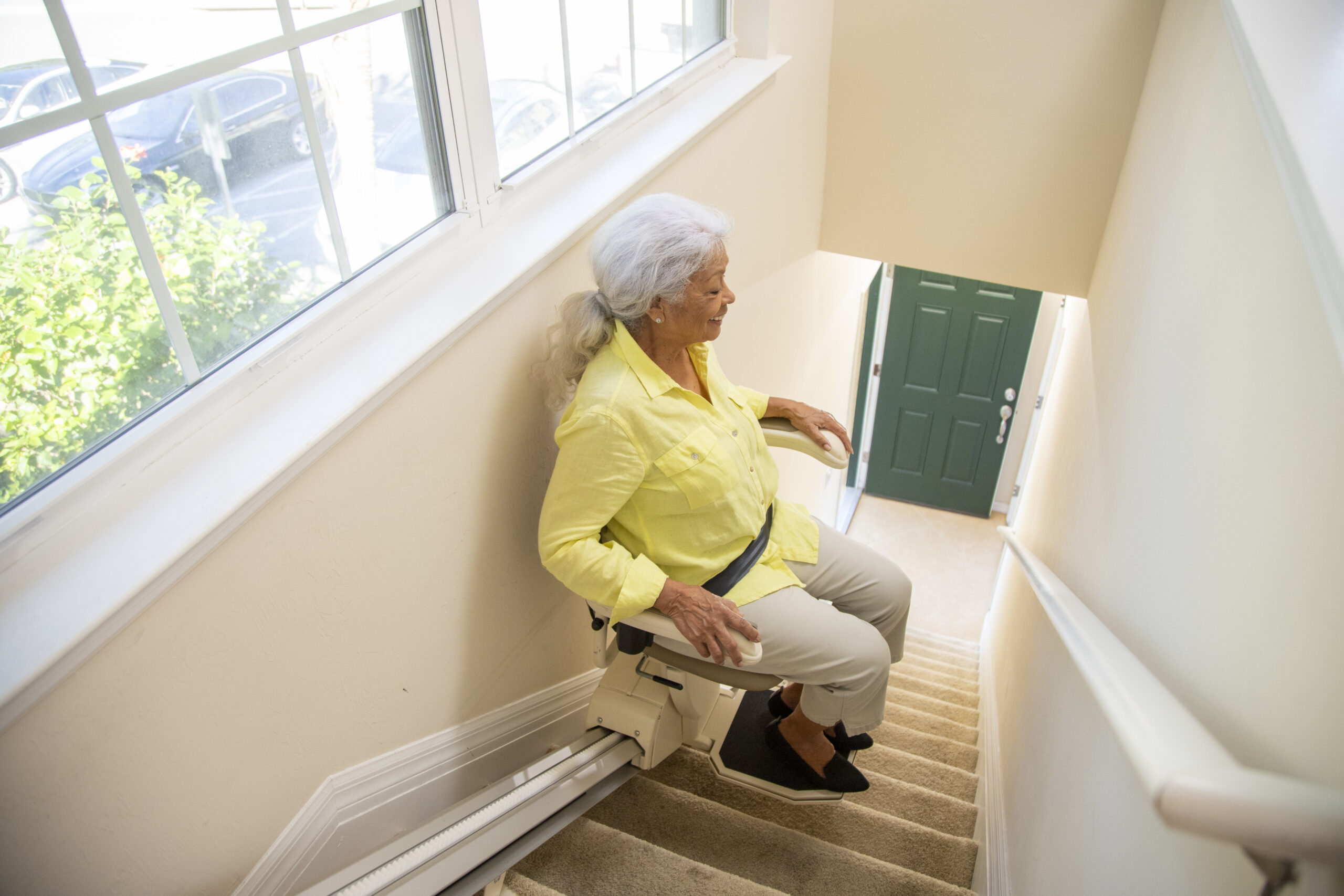 Older woman in a yellow shirt riding a straight stair lift along a carpeted staircase in her home.
