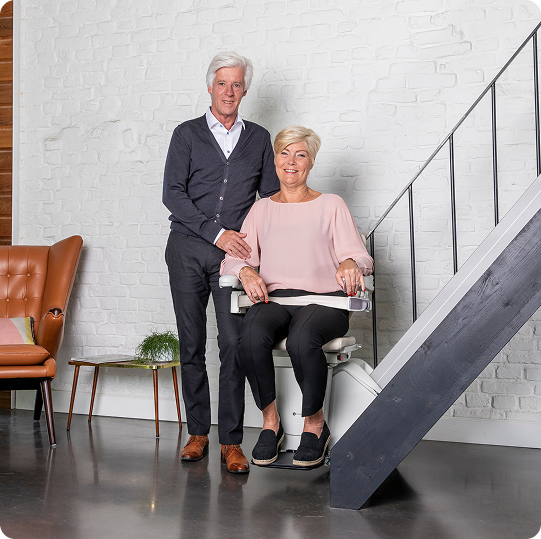 A man and woman facing the camera. The woman is wearing a pink shirt and sitting in a stair lift. The man is in a gray sweater and is holding her arm gently.
