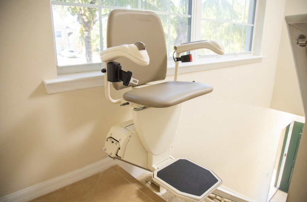 a stair lift at the top of stairs, with a window behind the lift looking out at trees. There is a green door at the bottom of the stairs.